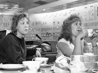 Schwarzweißfotografie von zwei Frauen, die in einem Raum voller Pläne und Schalter etwas aus Kaffeetassen trinken.