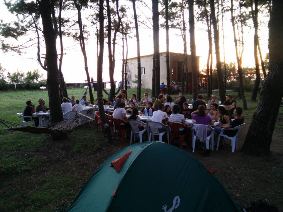 Eine Gruppe von etwa dreißig Leuten sitzt zwischen Bäumen auf Plastikstühlen im Abendlicht. Im Vordergrund ein grünes Zelt und eine Hängematte, im Hintergrund ein Haus mit Flachdach.