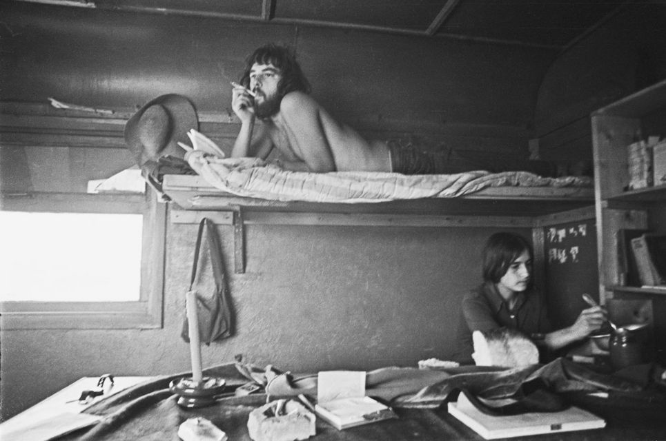 Schwarzweiß-Fotografie zeigt das Innere einer Art Wohnwagen mit Schlafebene, auf der ein Mann liegt und raucht, unter ihm sitzt eine junge Frau.