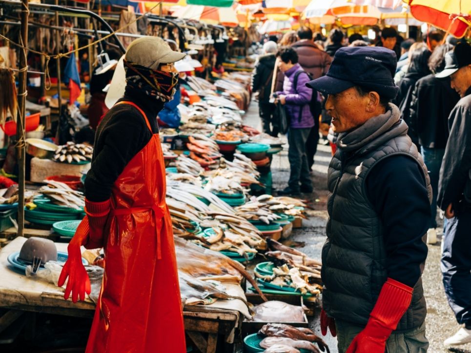 Stände mit Fellen und anderen Körperteilen von Tieren auf einem Markt, ein Mann steht einem Verkäufer gegenüber, der eine rote Schürze trägt und Mund und Nase mit einem Schal verdeckt.