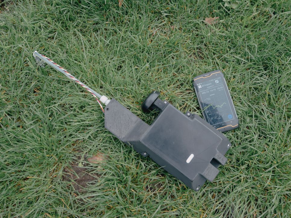 Das schwarze Messgerät mit einem Rohr, daneben ein Smartphone mit Schadstoff-Diagramm