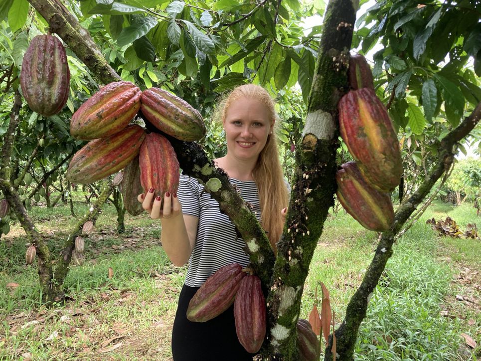Franziska Krause steht hinter einer Kakaopflanze, sie hält eine Kakaofrucht in der Hand.