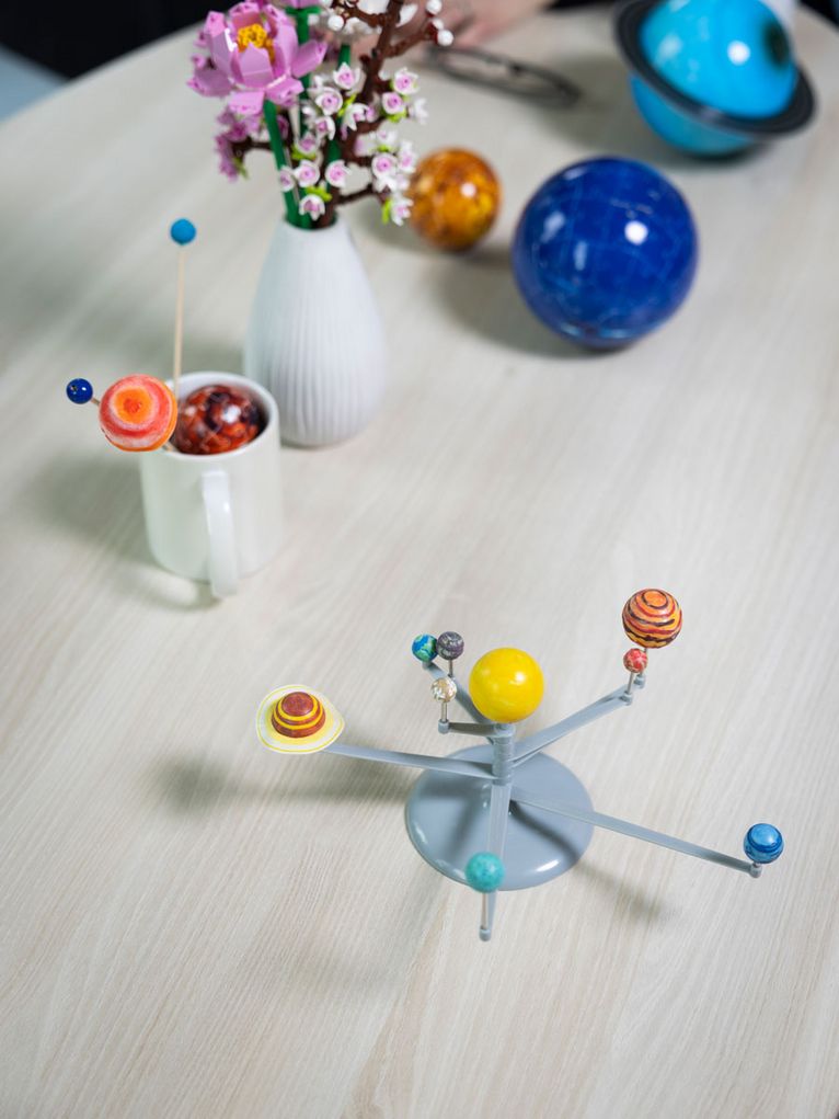 Auf einem Tisch stehen mehrere kleine Modelle von Planeten und Himmelskörpern, darunter ein mechanisches Modell des Sonnensystems. Im Hintergrund ist eine Blumenvase zu sehen, darin stehen aus Lego-Bausteinen geformte Plastikblumen.