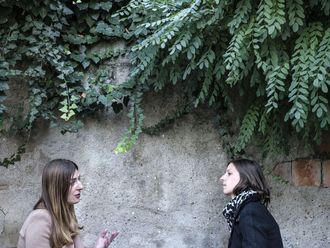 Zwei junge Frauen im Gespräch. Sie stehen vor einer grün bewachsenen Mauer.