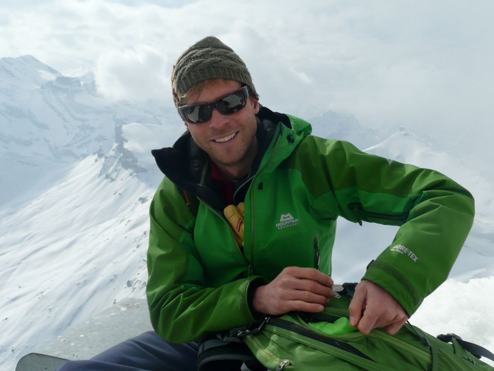 Porträt von Christian Reichel mit Mütze, Sonnenbrille und grüner Outdoor-Jacke vor verschneitem Bergpanorama.