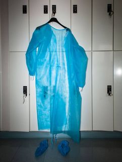 Ein blauer, leicht durchsichtiger medizinischer Schutzanzug hängt an einem Bügel an vor Schließfächern, darunter zwei blaue Überschuhe. Zusammen ähnelt es einer Person.