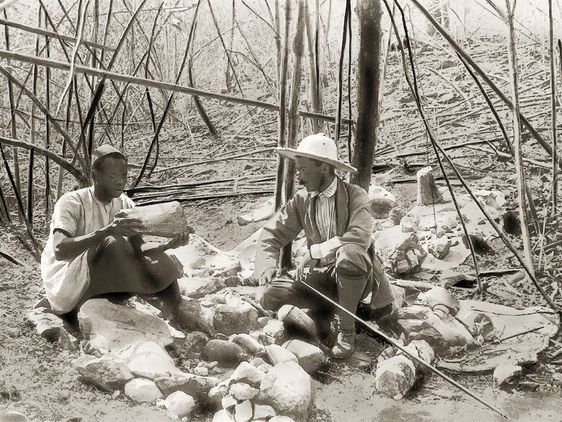 Schwarz-weiß Fotografie zeigt zwei Männer, die im Wald auf dem Boden inmitten von Knochenteilen sitzen. Einer hält ein großes Fragment in der Hand.