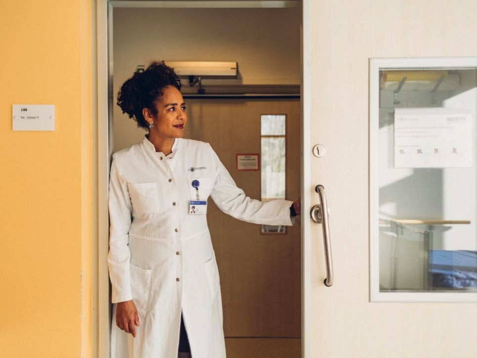 Marylyn Addo im Arztkittel im Krankenhausflur in einem Türrahmen.