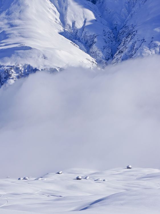 Schneebedeckte Berglandschaft, kleine verschneite Hütten, eine Nebelschicht durchzieht die ansonsten sonnige Landschaft.