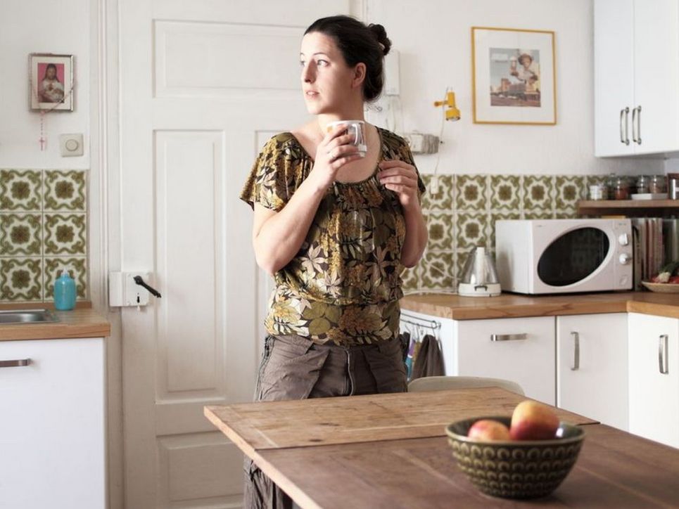 Eine Frau steht mit einer Tasse in der Hand in einer Küche, den Blick zur Seite gerichtet.