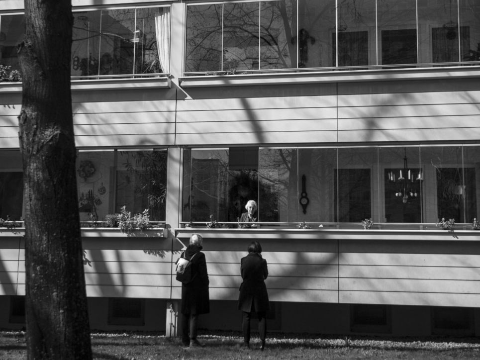 Zwei Personen vor einem Wohngebäude, sie sprechen mit einer älteren Frau, die sich auf dem Balkon befindet.