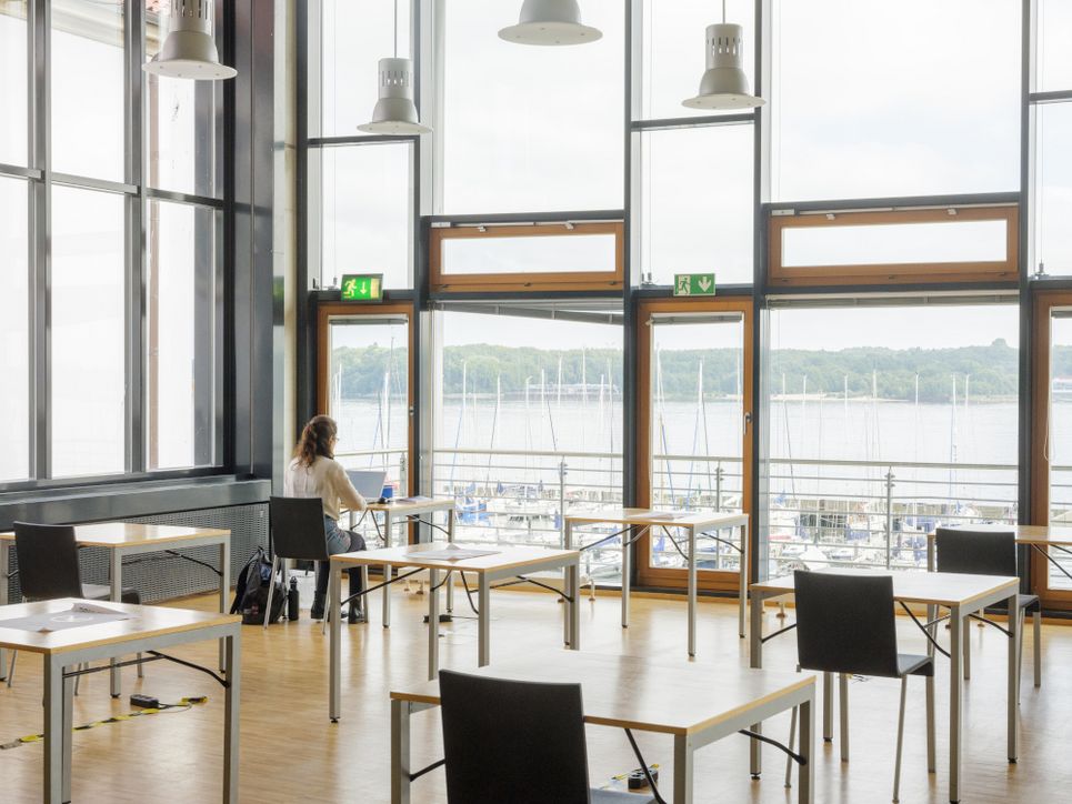 Lichtdurchfluteter Bibliotheksraum, rundum verglast und mit Blick aufs Wasser, eine Frau sitzt an einem der vorderen Tische.