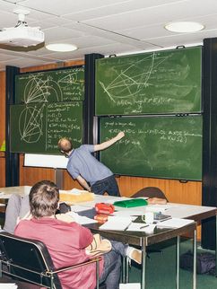 Mathematiker in einem Raum mit vollgeschriebener Tafel, an die jemand etwas schreibt.