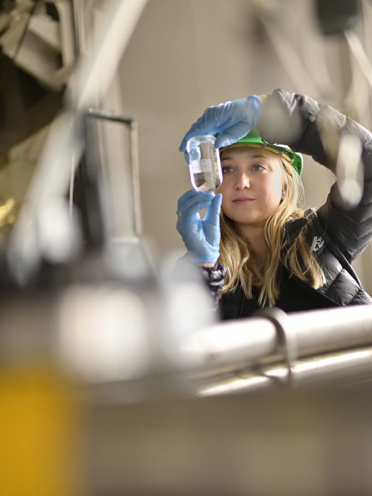 Eine Frau mit grünem Helm im Labor, die auf einen Glasbehälter schaut. 