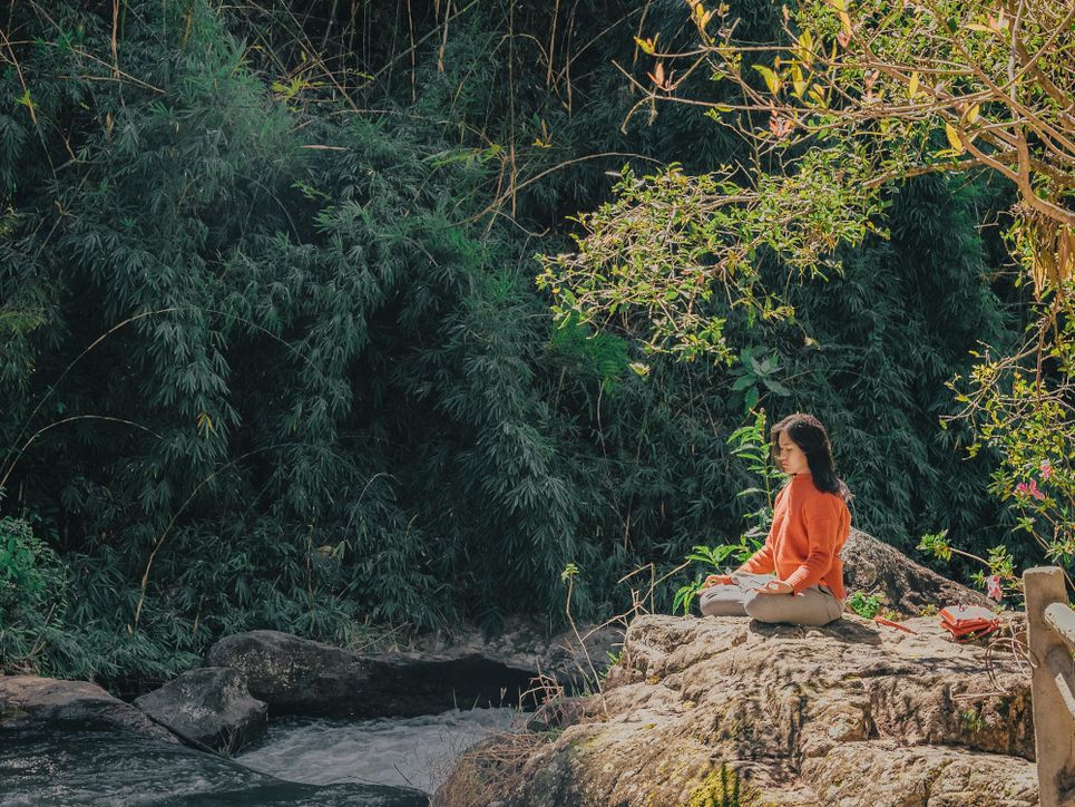 Eine Frau sitzt im Schneidersitz mitten in der Natur auf einem Felsen an einem Fluss und meditiert.