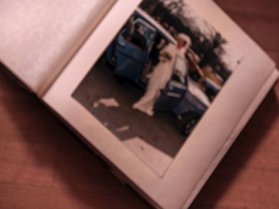 Ein geöffnetes Fotoalbum auf einem Holztisch. Auf dem Foto ist eine Frau vor einem Auto zu sehen. Das Foto ist verschwommen.