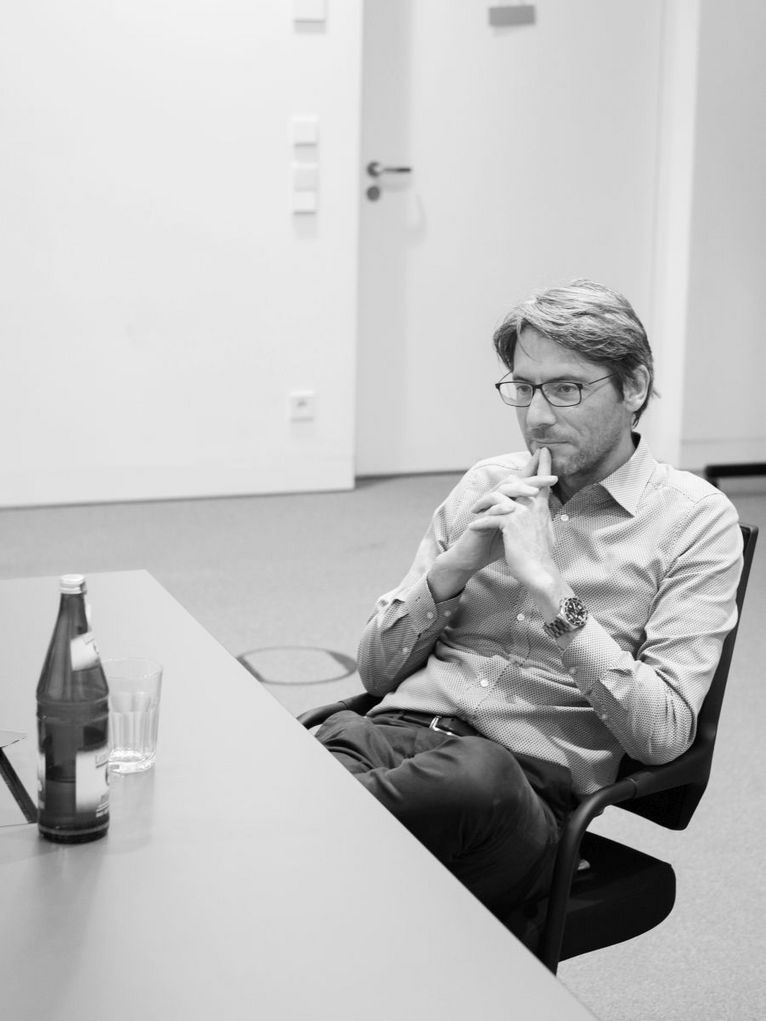 Schwarzweißporträt von Swen Schulz, der an einem Tisch sitzt, vor ihm eine Flasche und ein Wasserglas
