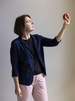 Portrait Prof. Dr. Olga Ramich Olga Ramich steht vor einer hellen Wand und hält einen roten Apfel in die Höhe. Sie blickt darauf und trägt ein dunkles Shirt, einen dunkelblauen Blazer sowie eine hellrosa Hose.