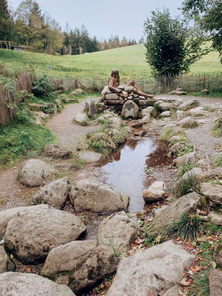 Wasser zwischen Steinen mit einer liegenden, langhaarigen Steinfigur, ein Weg führt darum herum.