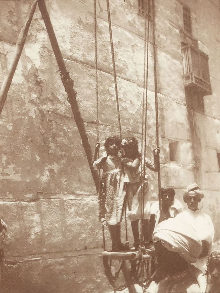 Historisches Foto zweier Mädchen auf einer Schaukel und andere Personen auf der Straße.