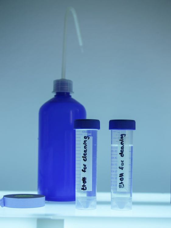 Blaue Flasche mit dünnem Ausguss sowie zwei beschriftete Reagenzgläser, auf denen steht: "E+OH for cleaning."