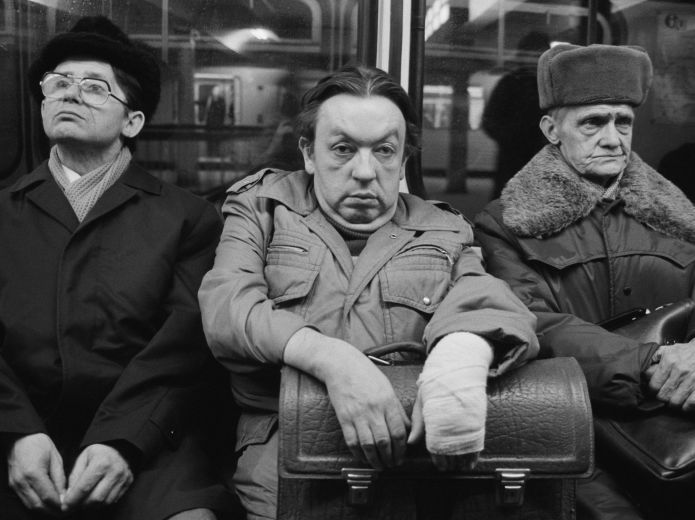Schwarz-Weiß Fotografie von drei Männern, die eng nebeneinander in einem öffentlichen Verkehrsmittel sitzen. Der mittlere schaut mit skeptischer Miene direkt in die Kamera.
