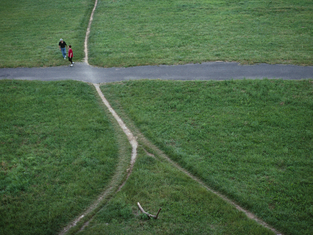 Von oben fotografierte, sich kreuzende Wege auf einer grünen Wiese, die in fünf Richtungen zeigen. Zwei Personen stehen an der Kreuzung.