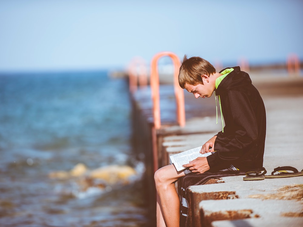 Ein Jugendlicher sitzt lesend am Meeresrand.