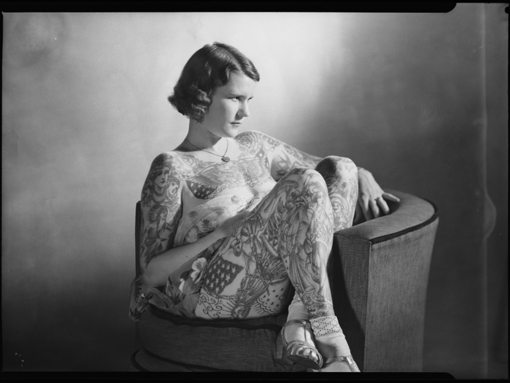 Die am ganzen Körper tätowierte Lady Betty Broadbent sitzt mit überschlagenen Beinen auf einem Sessel.